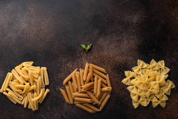 assorted uncooked pasta (treccia, farfalle, whole grain penne) on a dark background. assorted uncooked pasta (treccia, farfalle, whole grain penne pasta) on a dark background. food frame. flat lay. carbohydrate food type photos stock pictures, royalty-free photos & images