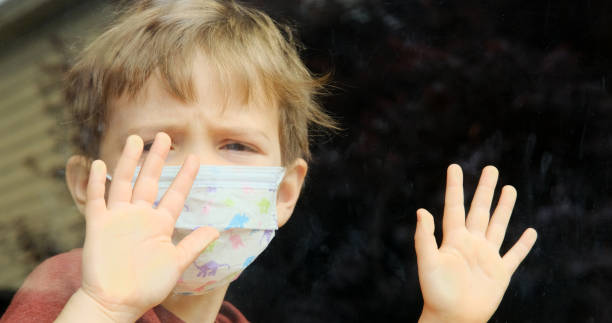 istock Little child boy looking through a window using a surgical mask due Covid-19 1218430078
