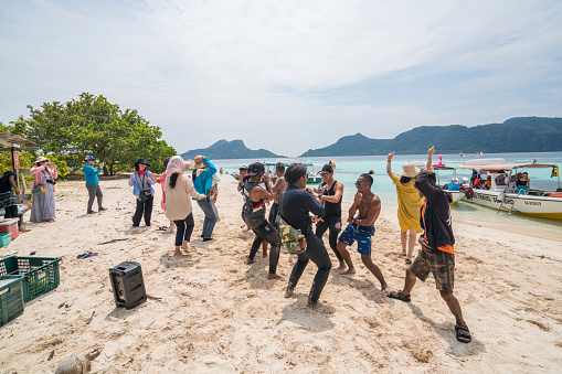 Semporna, Malaysia - November 6, 2019: On nice sunny day, Chinese tourists and local guides enjoying dancing on on sandy beach on small Pom Pom island near Semporna on Island Borneo, Malaysia. In background  are tourists hiding in shadow and taking photos.