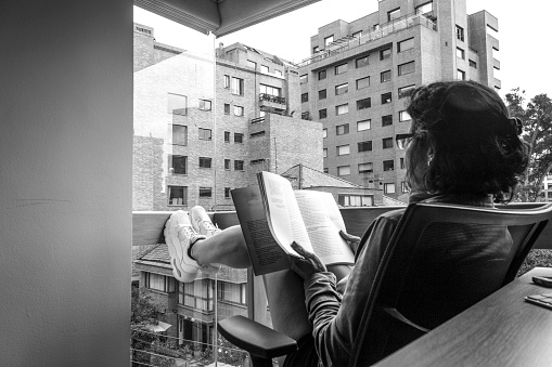 Girl reading under the light that enters through a panoramic window, where you can see the street, the buildings around and part of the room\nSeries of images about an activity carried out during social isolation\nBlack and White image