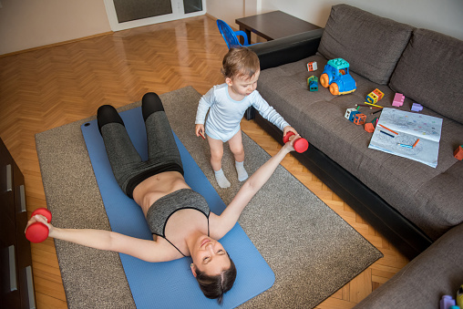 Young woman, stay at home mom, working out with dumbbells in a living room, lying down on a exercise mat, while her toddler son, baby boy, playing around with toys. Young family spending time together living healthy life, exercising at home, during coronavirus, covid 19 quarantine.