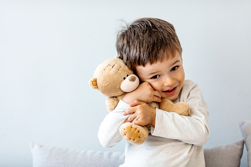 Portrait of one Happy toddler boy playing with his teddy bear at home in front of Gray wall.
