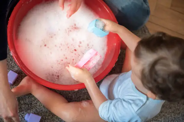 Toddler boy sitting on a floor in a living room and playing, washing and cleaning toys in a washbowl with his mother. Cleaning everything and doing chores in a coronavirus isolation and quarantine.