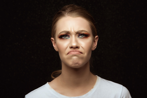 Blonde girl feeling angry and displeased making a face, blue eyes and strange makeup wearing white T-shirt posing on black background