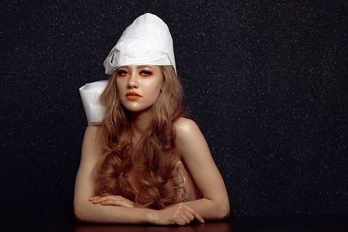 Blond girl posing in studio on black background with hat out of toilet paper