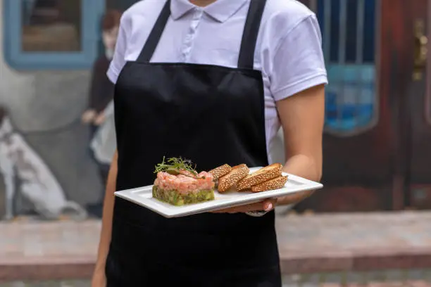 a girl in a black apron and a white bozka is holding in front of her a salmon avocado and rucola appetizer, 
white bread cracker on a white plate. background street