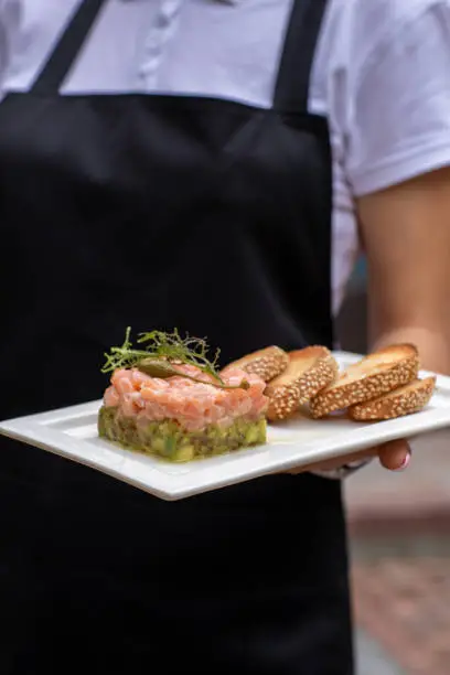 a girl in a black apron and a white bozka is holding in front of her a salmon avocado and rucola appetizer, 
white bread cracker on a white plate. background street