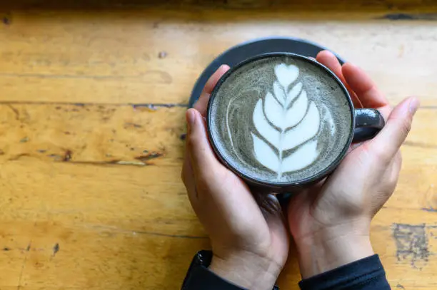 Photo of Someone hands holding a cup of Goth Latte or Charcoal Latte on wooden table (Focus on coffee surface).