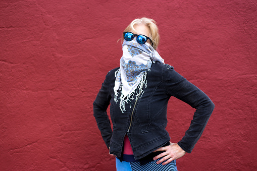 A woman wearing a bandana/scarf mask stands with arms crossed against a red background. Shot in Santa Fe, NM.
