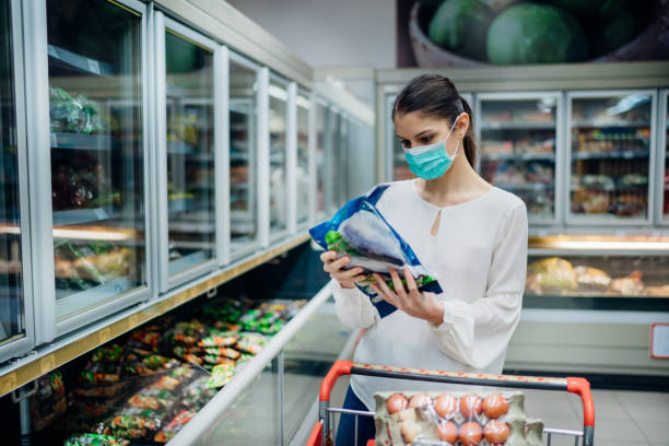 Woman with hygienic mask shopping for supplies.Pandemic quarantine preparation.Choosing nonperishable food essentials from store shelves.Budget buying at a supply store.Food supplies shortage. stock photo