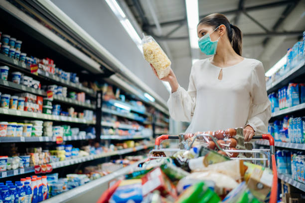Young person with protective face mask buying groceries/supplies in the supermarket.Preparation for a pandemic quarantine due to coronavirus covid-19 outbreak.Choosing nonperishable food essentials stock photo