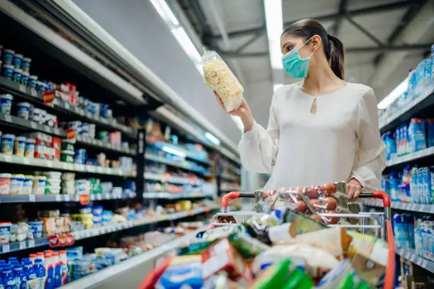 Photo of Young person with protective face mask buying groceries/supplies in the supermarket.Preparation for a pandemic quarantine due to coronavirus covid-19 outbreak.Choosing nonperishable food essentials