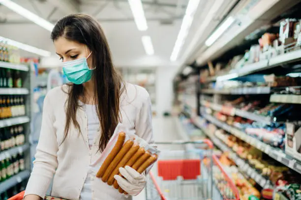 Woman with protective mask and gloves shopping for supply.Budget shopping at a supermarket.Buying processed nonperishable food.Preparation for a pandemic quarantine due to covid-19.Expiration date