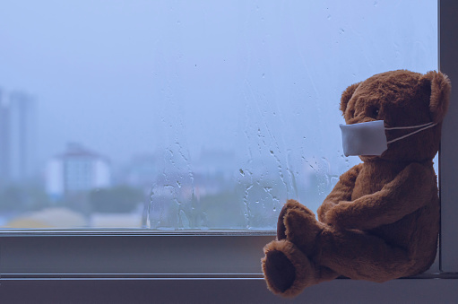 Teddy bear wearing mask sitting at window while raining in monsoon season. Stay home away from virus concept.