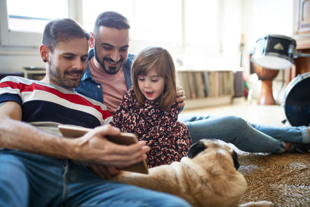 Relaxed Fathers and 3 Year Old Daughter Using Digital Tablet Smiling Spanish fathers enjoying weekend leisure with young daughter and dog as they use digital tablet together on living room floor. gay daddy stock pictures, royalty-free photos & images