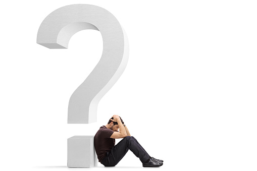 Sad young man sitting on the floor holding his head and leaning against a big question mark isolated on white background