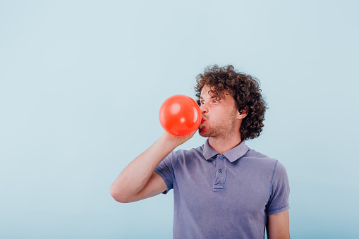 Grimacing unshaven man with curly hair in blue shirt blowing red balloon on blue studio background