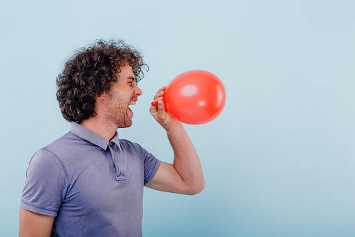 Side view of excited casual curly man closing eyes and laughing while inhaling helium gas from balloon having funny voice
