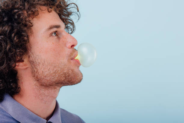 Portrait of a young man squinting while blowing a bubble from chewing gum and looking at camera.