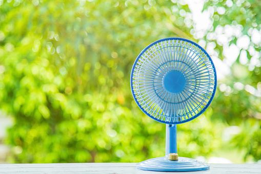Close up blue plastic desktop electric fan on white wooden table with green blurred nature background with blank left side for copy space.