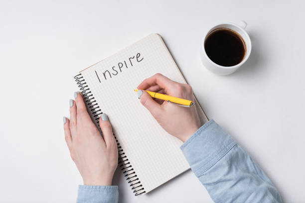 istock Female hand writes words Inspire in copybook. Notepad, pen and coffee on white background. 1218539869