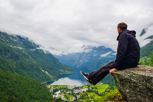 Male tourist sitting on the cliff edge near Flydalsjuvet Viewpoint. The summer landscape of Geiranger small village which is located at the end of the Geirangerfjord. Norway.