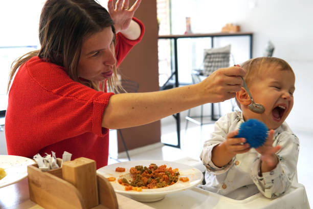 unhappy child refusing to eat at a restaurant with white background, while single mom is trying to deceive the toddler by tricks; anyway he does not want to eat