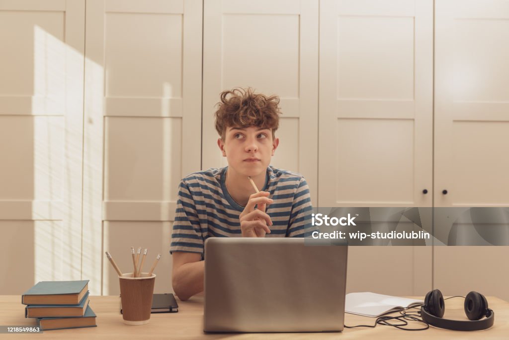 Miły nastoletni chłopiec w słuchawkach korzysta z laptopa i uczy się przy biurku w swoim pokoju. Robi notatki ołówkiem. Uczenie się na odległość z powodu epidemii koronawirusa - Zbiór zdjęć royalty-free (Choroba zakaźna)