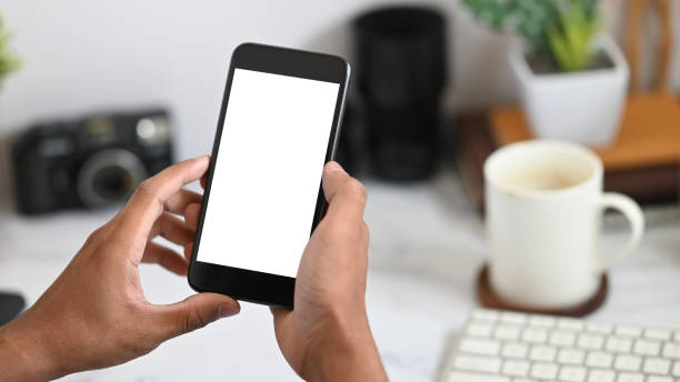 istock Cropped image of hands holding a white blank screen smartphone over keyboard, coffee cup and office equipment as background. Orderly workspace concept. 1218554513