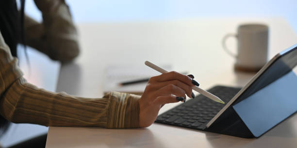 istock Cropped image of creative woman's hands holding a stylus pen and using a computer tablet with keyboard case while sitting at the white working desk over comfortable office as background. 1218554592