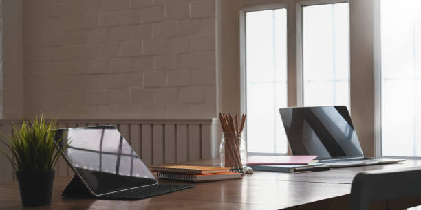 istock Comfortable living room. Orderly workplace. Computer tablet,laptop putting on wooden working table with pencil holder, potted plant and stack of notebook over living room as background. 1218554748