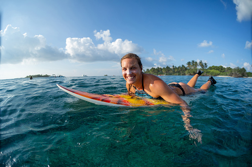 Happy woman surfer lies on the surf board in the ocean in Maldives. Girl looks at the camera and smiles