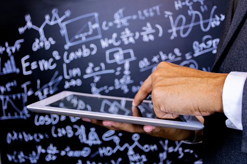 Close to the hand of the teacher using a tablet, Background is blackboard with a lot of mathematical formulas, The characters are glowing.