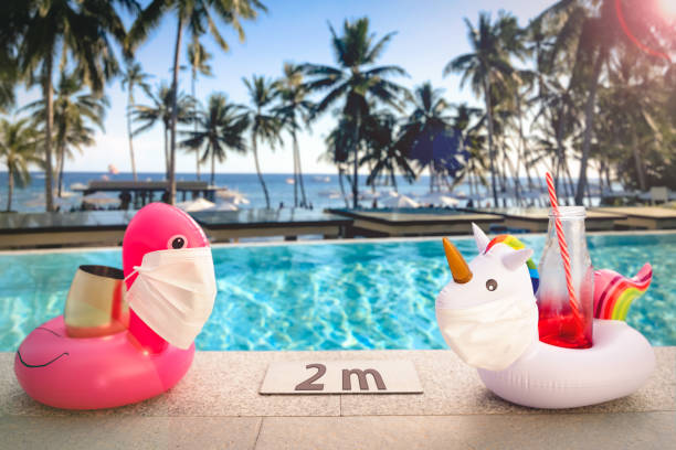 надувные игрушки у бассейна в тропическом курортном бассейне в масках для лица, находящихся в 2 метрах от отеля, чтобы сохранить социальную  - aside стоковые фото и изображения