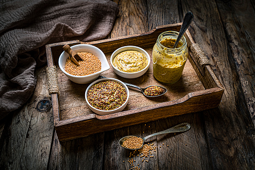 Spices: two bowls filled with whole grain and Dijon mustard shot on dark wooden table. The third bowl is filled with dried mustard seeds. Spoons with mustard are beside the bowls. Predominant colors are gold and brown. High resolution 42Mp studio digital capture taken with Sony A7rII and Sony FE 90mm f2.8 macro G OSS lens