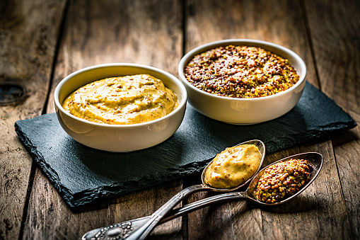 Spices: close up view of two bowls filled with whole grain and Dijon mustard shot on dark wooden table. Spoons with mustard are in front of the bowls. Predominant colors are gold and brown. High resolution 42Mp studio digital capture taken with Sony A7rII and Sony FE 90mm f2.8 macro G OSS lens