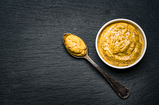 Spices: fresh organic Dijon mustard in a white bowl shot from above on dark slate background. A spoon with mustard is beside the bowl. The composition is at the right of an horizontal frame leaving useful copy space for text and/or logo at the left. Predominant colors are gold and black. High resolution 42Mp studio digital capture taken with Sony A7rII and Sony FE 90mm f2.8 macro G OSS lens