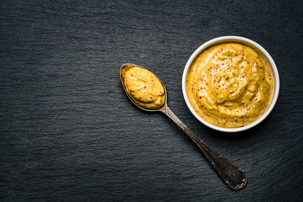 Dijon mustard in a bowl Spices: fresh organic Dijon mustard in a white bowl shot from above on dark slate background. A spoon with mustard is beside the bowl. The composition is at the right of an horizontal frame leaving useful copy space for text and/or logo at the left. Predominant colors are gold and black. High resolution 42Mp studio digital capture taken with Sony A7rII and Sony FE 90mm f2.8 macro G OSS lens dijon stock pictures, royalty-free photos & images