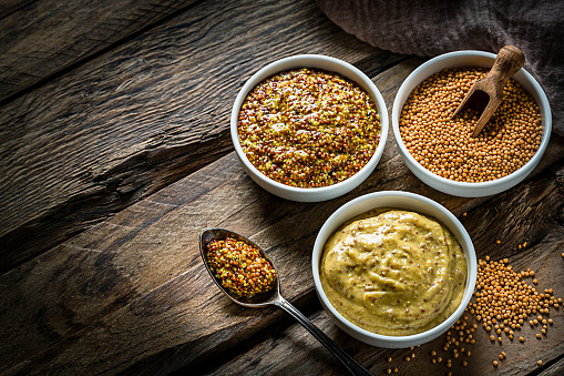 Spices: two bowls filled with whole grain and Dijon mustard shot from above on dark wooden table. The third bowl is filled with dried mustard seeds. Spoons with mustard are beside the bowls. The composition is at the right of an horizontal frame leaving useful copy space for text and/or logo at the left. Predominant colors are gold and brown. High resolution 42Mp studio digital capture taken with Sony A7rII and Sony FE 90mm f2.8 macro G OSS lens