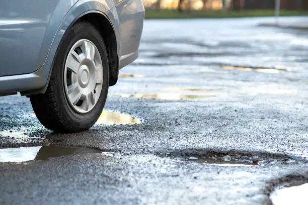 Close up of car wheel on a road in very bad condition with big potholes full of dirty rain water pools.