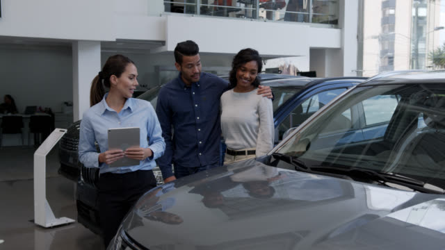 Saleswoman holding a tablet showing cars to a happy diverse couple at the dealership