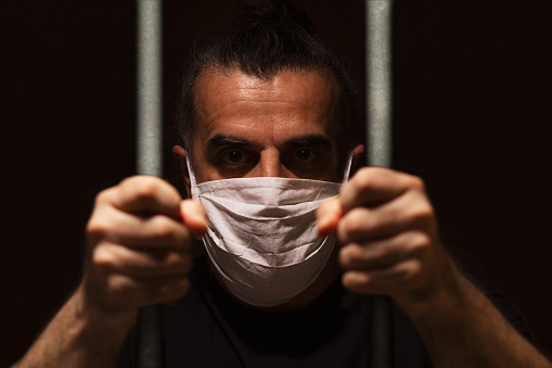 Hombre con máscara médica blanca de pie en la oscuridad, sosteniendo barras con sus propias manos, encerrado en la oscuridad, en aislamiento y cuarentena photo