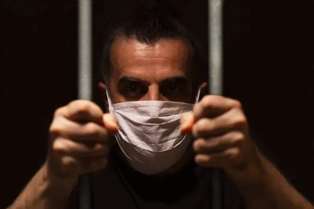 Man with white medical mask standing in the dark, holding bars with his bare hands, locked away in dark, in isolation and quarantine