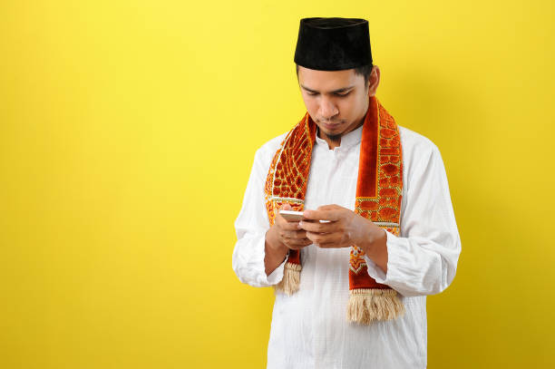 Young Asian Muslim man wearing Muslim clothes holding mobile phone look at phone screen Young Asian Muslim man wearing Muslim clothes holding mobile phone look at phone screen, isolated on yellow background 3381 stock pictures, royalty-free photos & images