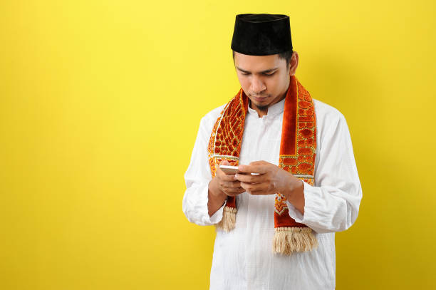 jovem muçulmano asiático vestindo roupas muçulmanas segurando olhar celular na tela do telefone - 3381 - fotografias e filmes do acervo