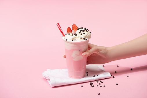 Cropped view of female hand with disposable cup of milkshake with chocolate morsels and strawberry on napkins on pink