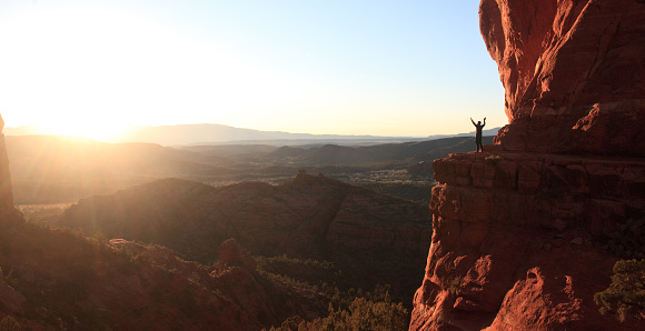 A male hiker - back view - in the beautiful red rocks of Sedona, Arizona. Hiker is in silhouette near sunset at the dramatic Cathedral Rock summit lookout.
