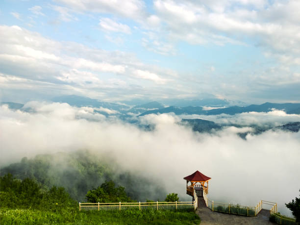 From above of beautiful viewing gazebo located on covered with green plants top of mountain with fog and cloudy sky in background