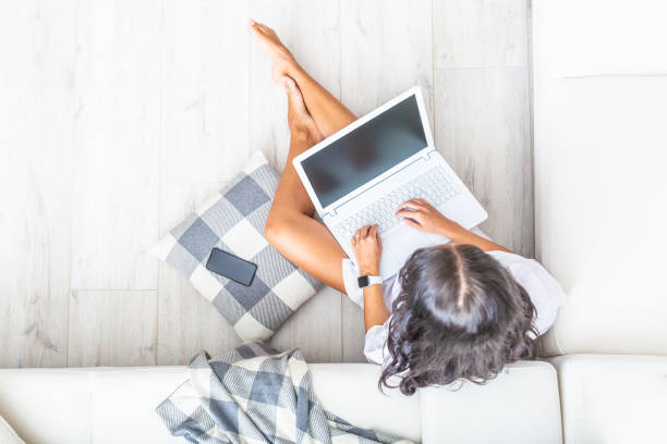 istock Top view of home office woman, working on a notebook, leaned against a white sofa, looking into the pc, bare legs, her cell phone next to her on a grey and white checked pillow. 1218626429