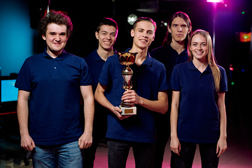 Group of young gamers in blue tshirts standing in computer club, team leader holding golden cup, they winning in cybersports competition